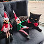 cat, black_cat, red_bow_tie, elf_doll, holiday_decoration, chair, indoor, window, wooden_floor, toy, festive, christmas, plush_toy, decor, pet, feline, relaxing, holiday_season, cute, cozy