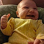 baby, smiling, thumbs_up, yellow_onesie, polka_dot, cushion, indoor, happy, infant, cute, child, portrait, closeup, soft_lighting, seated, clothing, person, young_child, expression, face