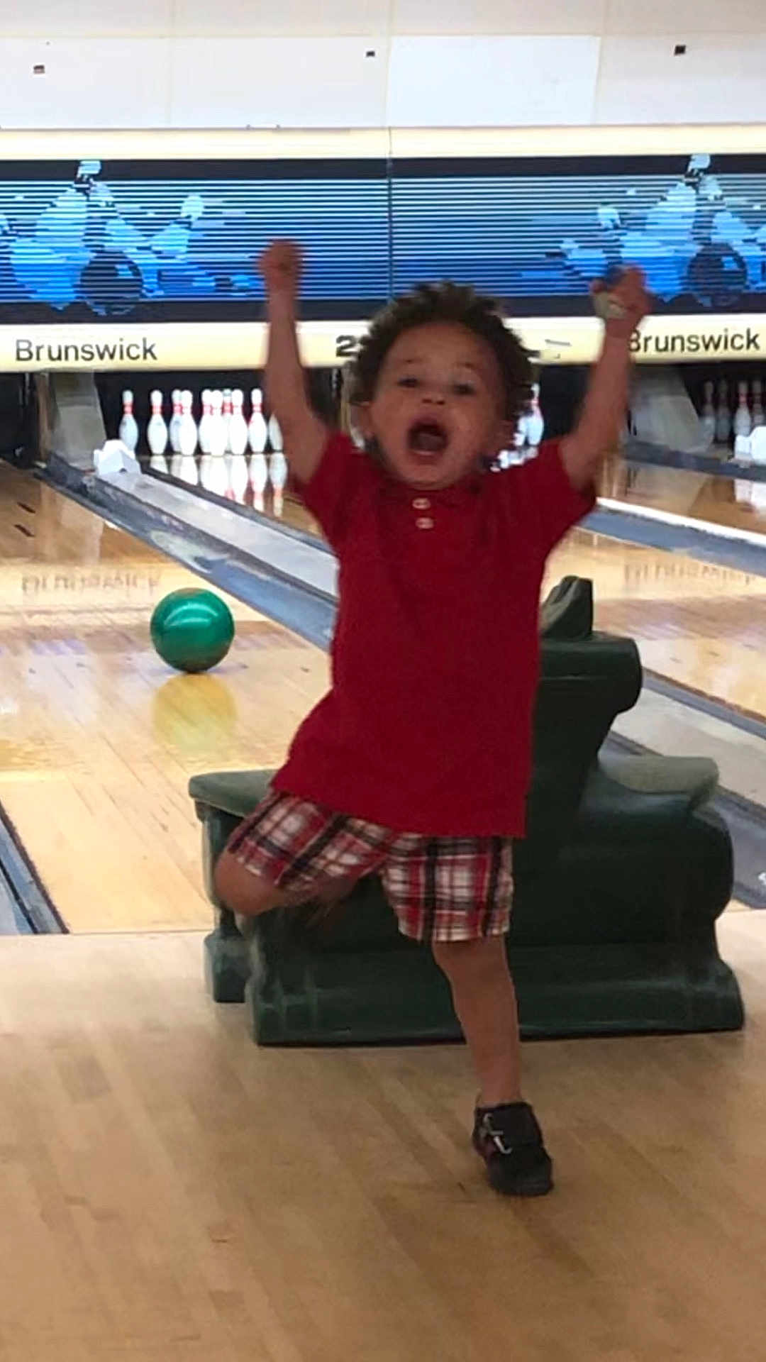 Michael is registered to the contest to win money with this photo: child, bowling_alley, bowling_ball, celebration, victory_pose, red_shirt, plaid_shorts, indoor, wood_floor, lane, pins, sport, happy, joy, person, active, motion, recreation, casual_clothing, fun