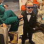 child, boy, tuxedo, sunglasses, smile, indoor, carpet, furniture, person, adult, hand_gesture, living_room, decor, bag, shoe, table, glass, plant, braided_hair, playful