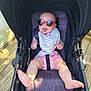 baby, sunglasses, stroller, infant, tongue_out, heart_shaped_glasses, pink_shorts, bib, sunlight, outdoor, wooden_deck, baby_boy, baby_girl, seat_belt, happy, cute, child, summer, smiling, fun