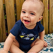 Brycen is registered to the contest to win money with this photo: baby, child, smiling, sitting, onesie, dinosaur_print, cushion, floral_pattern, wooden_fence, outdoor, happy, cute, infant, portrait, nature, plants, pink_flowers, barefoot, skin, head