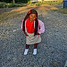 child, girl, portrait, outdoor, standing, braids, red_shirt, pink_jacket, skirt, sneakers, gravel_driveway, house, neighborhood, mailbox, trash_bin, trees, sunlight, fashion, serious_expression, full_body