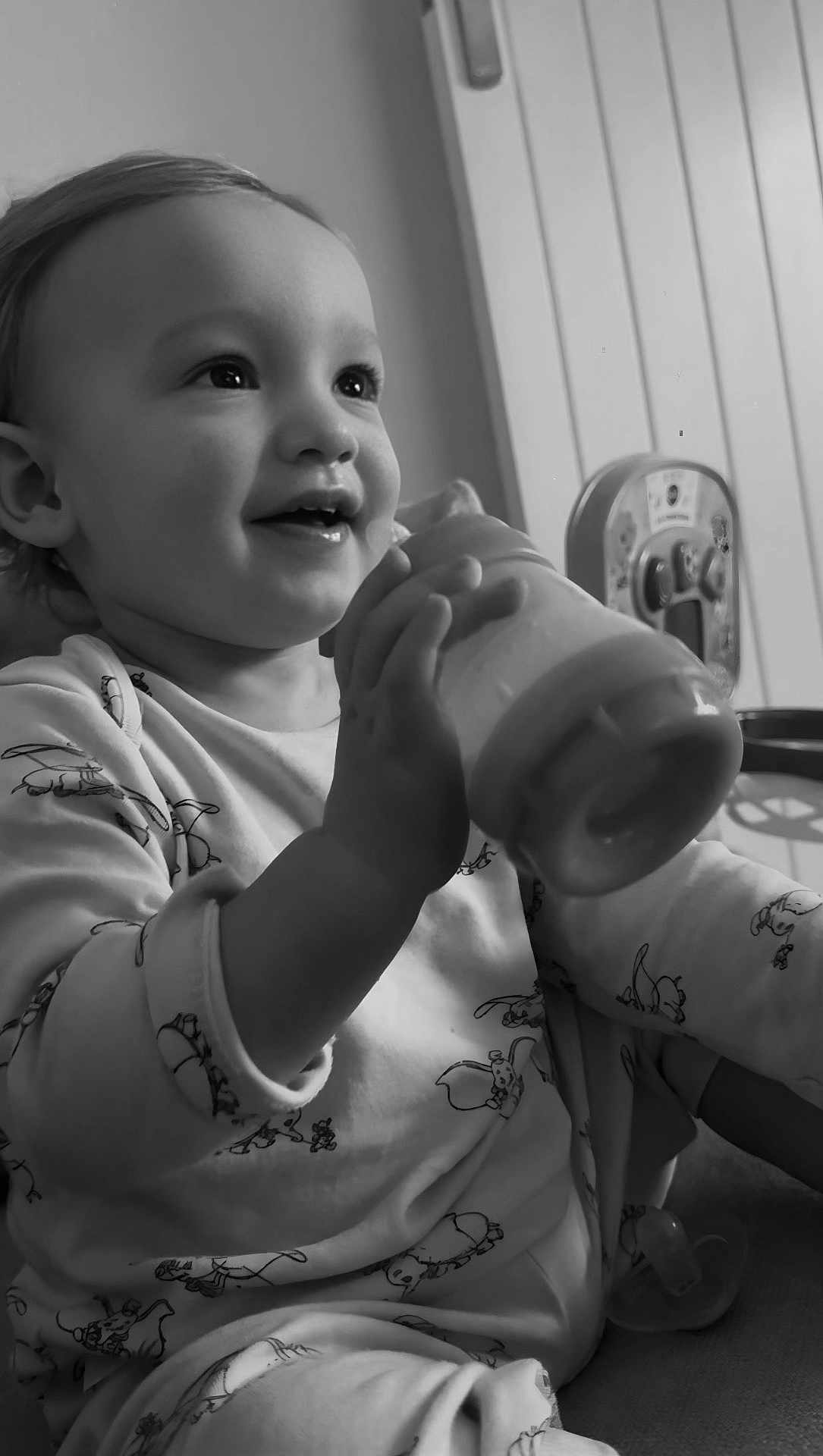 Luca is registered to the contest to win money with this photo: toddler, child, baby, smiling, bottle, pajamas, indoor, face, hand, sitting, happy, cute, feeding, young_child, portrait, monochrome, black_and_white, expression, cozy, person