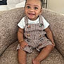 toddler, child, smile, happy, curly_hair, overalls, checkered, armchair, indoors, person, barefoot, bracelet, cute, portrait, seated, infant, clothing, furniture, baby, cheerful
