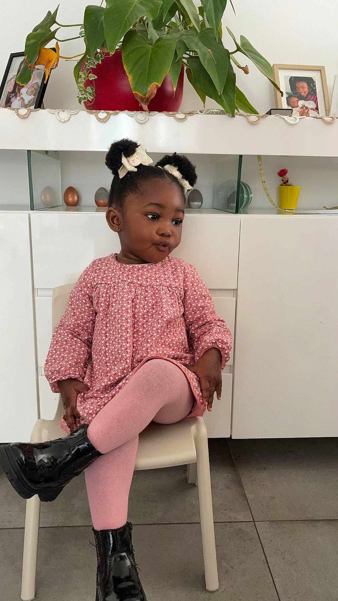 Aëlla a rejoint le concours — aidez-le/la à gagner de superbes lots ! child, toddler, girl, pink_dress, tights, black_boots, hair_bows, sitting, chair, white_cabinet, potted_plant, photo_frame, interior, floor_tiles, portrait, expression, cute, fashion, smile, home