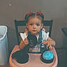 child, toddler, girl, portrait, sitting, high_chair, indoors, home, table, toy, glowing_ball, black_disc, red_bows, overalls, white_shirt, smiling, face, hair, chair, playtime