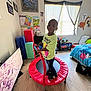 child, trampoline, bedroom, toys, posters, window, bed, floor, shoes, clothing, light, wall, furniture, flooring, education, play, indoor, person, young, colorful
