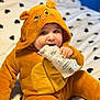 baby, child, costume, bear, hood, bed, bedspread, heart_pattern, cute, fuzzy, chewing, plastic_package, sitting, indoor, soft, clothing, person, infant, adorable, blue_eyes