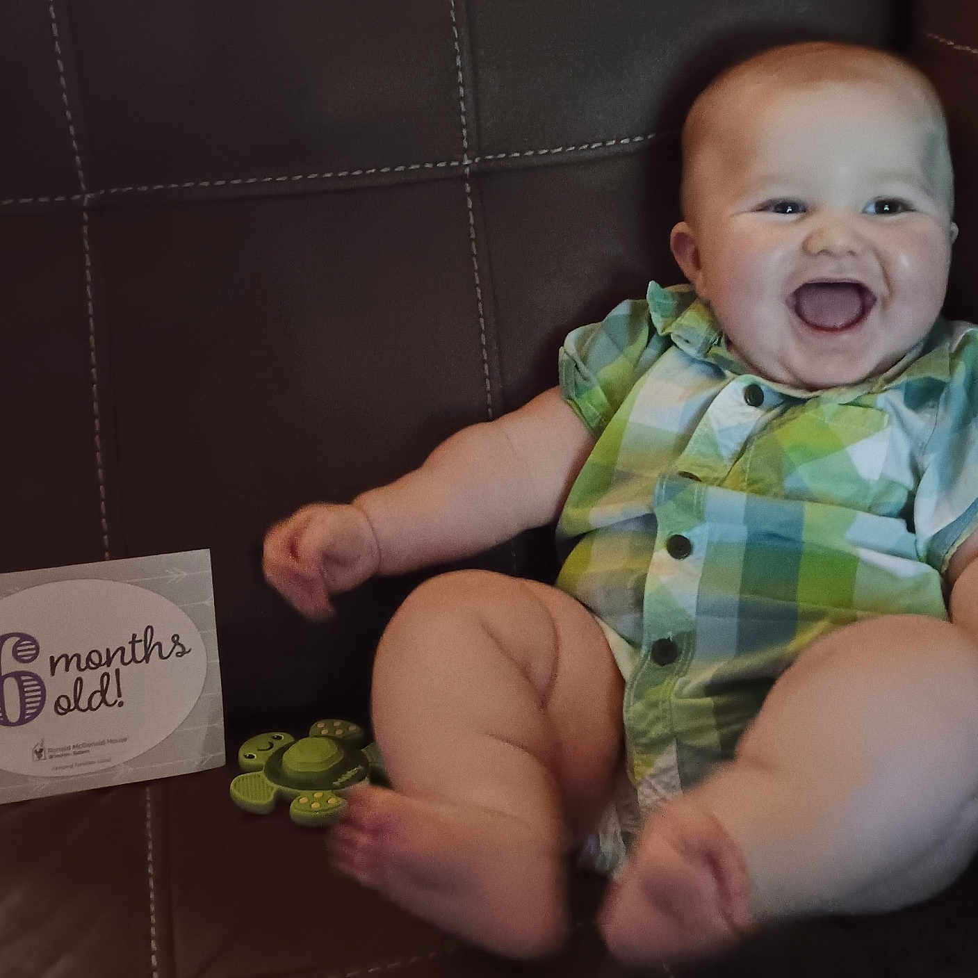 Jax joined the competition — help win amazing prizes! arms, baby, blue_clothing, child, couch, cute, face, green_clothing, happy, indoor, infant, laughing, leather, legs, plaid_shirt, sign, sitting, six_months, smiling, toy
