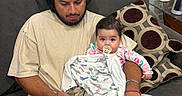 Gianna is registered to the contest to win money with this photo: baby, man, pacifier, blanket, couch, cushion, cap, phone, bracelet, ring, seahorse_statue, glass_table, indoor, person, home, casual_clothing, beard, sitting, looking_down, wooden_floor