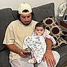 baby, man, pacifier, blanket, couch, cushion, cap, phone, bracelet, ring, seahorse_statue, glass_table, indoor, person, home, casual_clothing, beard, sitting, looking_down, wooden_floor