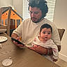 Gianna joined the competition — help win amazing prizes! baby, man, indoor, table, phone, curly_hair, sunglasses, baby_clothes, wooden_chair, toy_house, window_blinds, tabletop, person, child, facial_hair, curious, seated, casual_clothing, home, parenting