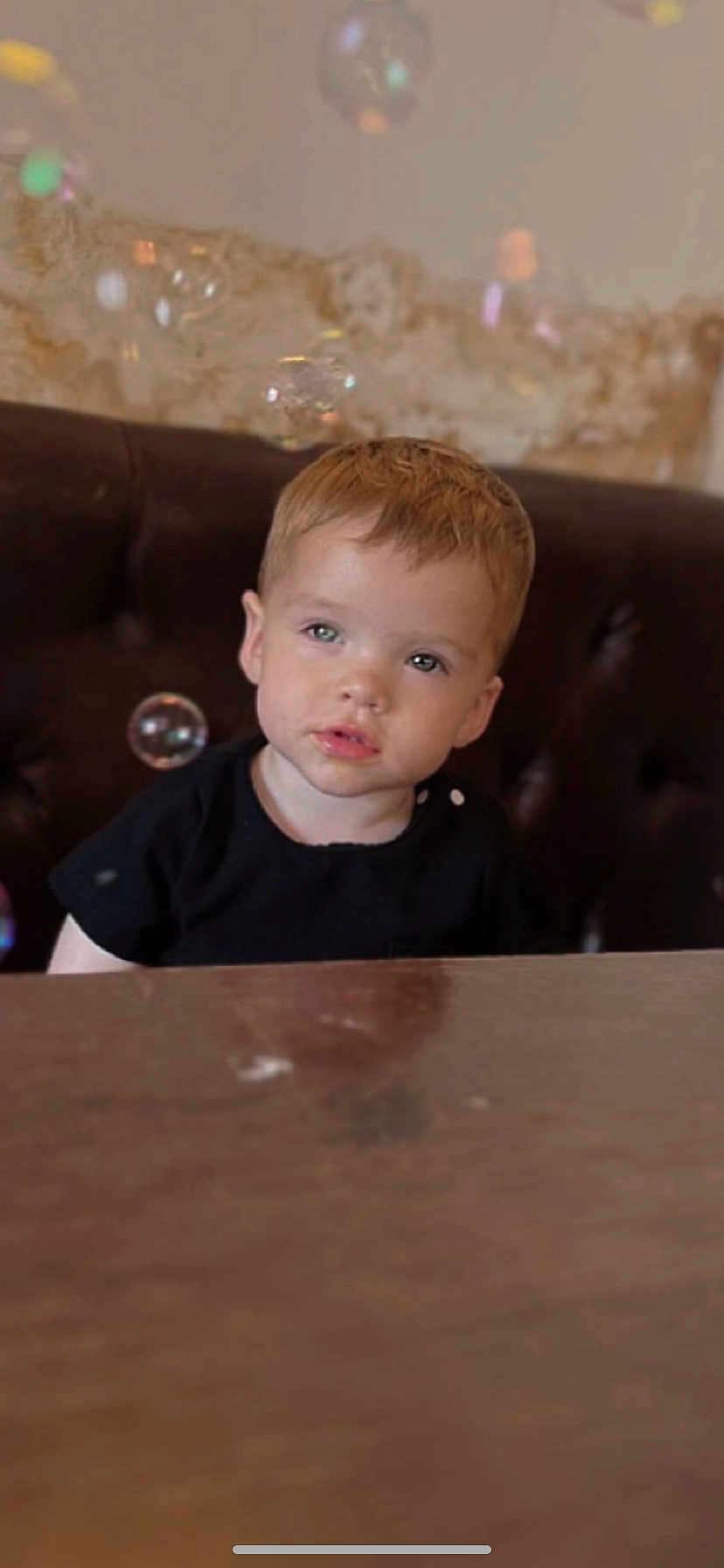 Elijah is registered to the contest to win money with this photo: child, toddler, boy, face, bubbles, table, indoor, curious, light_hair, black_shirt, seated, cushion, wooden_table, portrait, expression, young_child, bubble, casual_clothing, home, cute