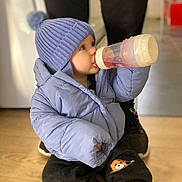 Elijah joined the competition — help win amazing prizes! toddler, child, baby_bottle, blue_jacket, knit_hat, pom_pom, indoor, wooden_floor, sitting, cozy_clothing, black_pants, cute, person, warm_clothes, floor, footwear, toy_design, baby, drinking, casual