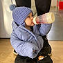 toddler, child, baby_bottle, blue_jacket, knit_hat, pom_pom, indoor, wooden_floor, sitting, cozy_clothing, black_pants, cute, person, warm_clothes, floor, footwear, toy_design, baby, drinking, casual
