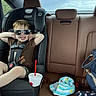 Levi is registered to the contest to win money with this photo: toddler, car_seat, sunglasses, drink_cup, car_interior, child, seatbelt, brown_shirt, shorts, happy, smiling, leather_seat, backseat, blue_bag, striped_clothing, sunlight, casual, relaxed, vehicle, window