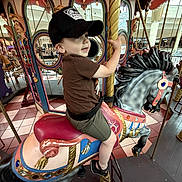 Levi is registered to the contest to win money with this photo: child, boy, carousel, carousel_horse, amusement_ride, indoor, cap, brown_shirt, shorts, sandals, mirror, colorful, playful, smiling, holding, pole, mall, floor_tiles, decorative, fun
