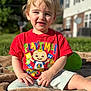 child, toddler, blond_hair, red_shirt, playtime, outdoor, grass, blanket, sunlight, smiling, sitting, shorts, daytime, house, background, cartoon, colorful, happy, person, portrait