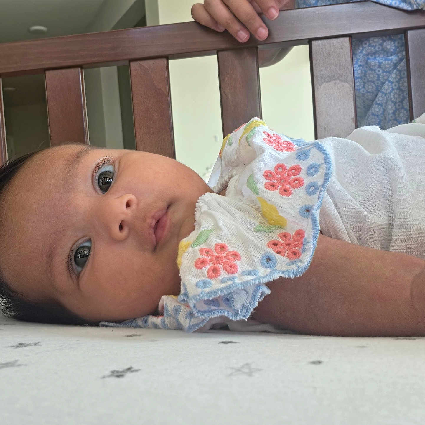Krishni is registered to the contest to win money with this photo: baby, bed, child, clothing, crib, curious, cute, embroidered, eyes, face, flower_pattern, hand, indoor, infant, lying_down, peaceful, person, portrait, sheet, wooden_rail