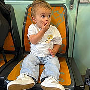 Louis a rejoint le concours — aidez-le/la à gagner de superbes lots ! toddler, child, seat, orange_seat, white_tshirt, jeans, sneakers, hand_on_mouth, surprised_expression, public_transport, window, green_wall, indoor, person, young_child, casual_clothing, seat_back, curly_hair, footwear, sitting