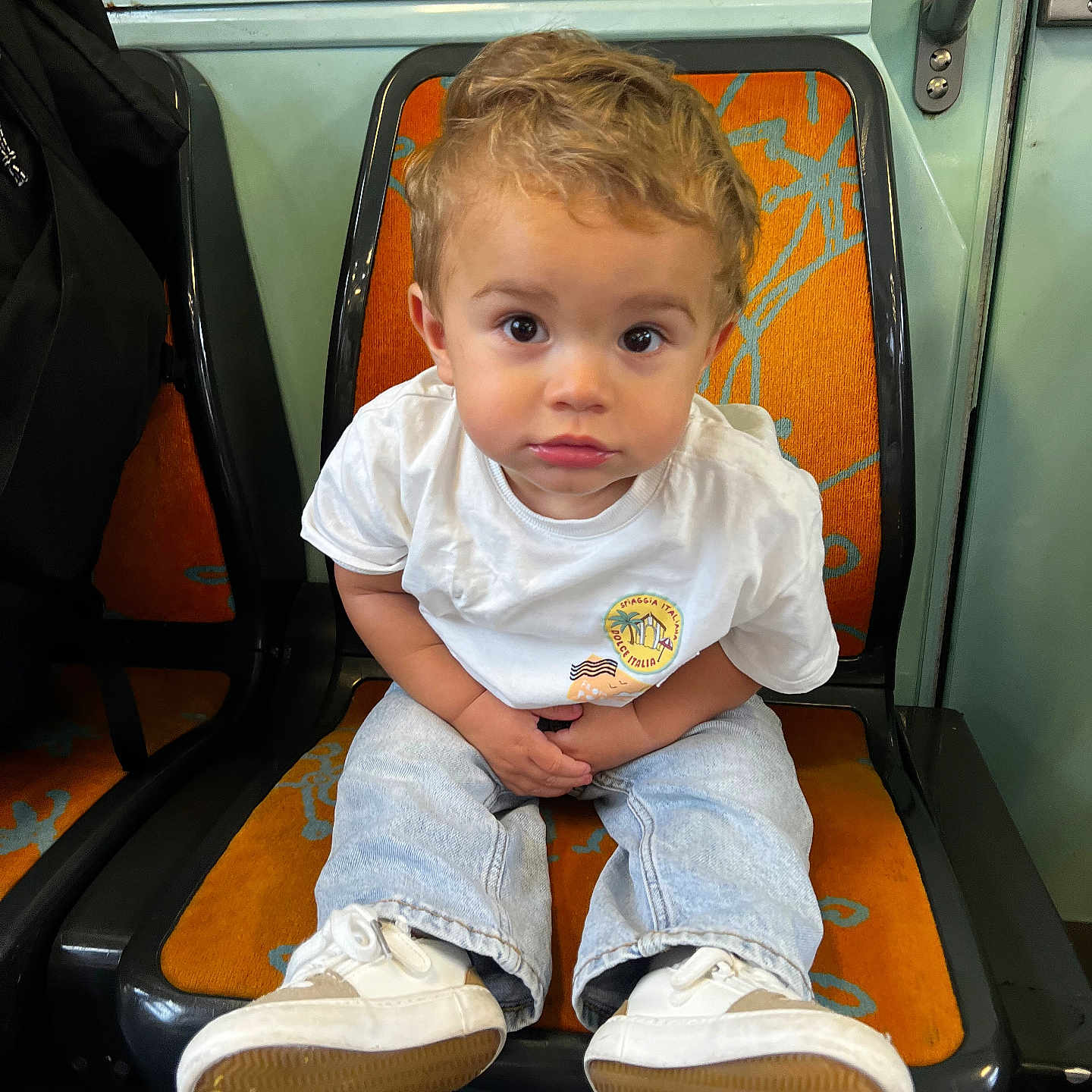 Louis participe au concours pour gagner de l'argent avec cette photo : casual_clothing, child, curly_hair, expression, face, floor, hands, indoor, jeans, looking_at_camera, orange_seat, person, public_transport, seat, shoes, sitting, toddler, transport, white_tshirt, young_child