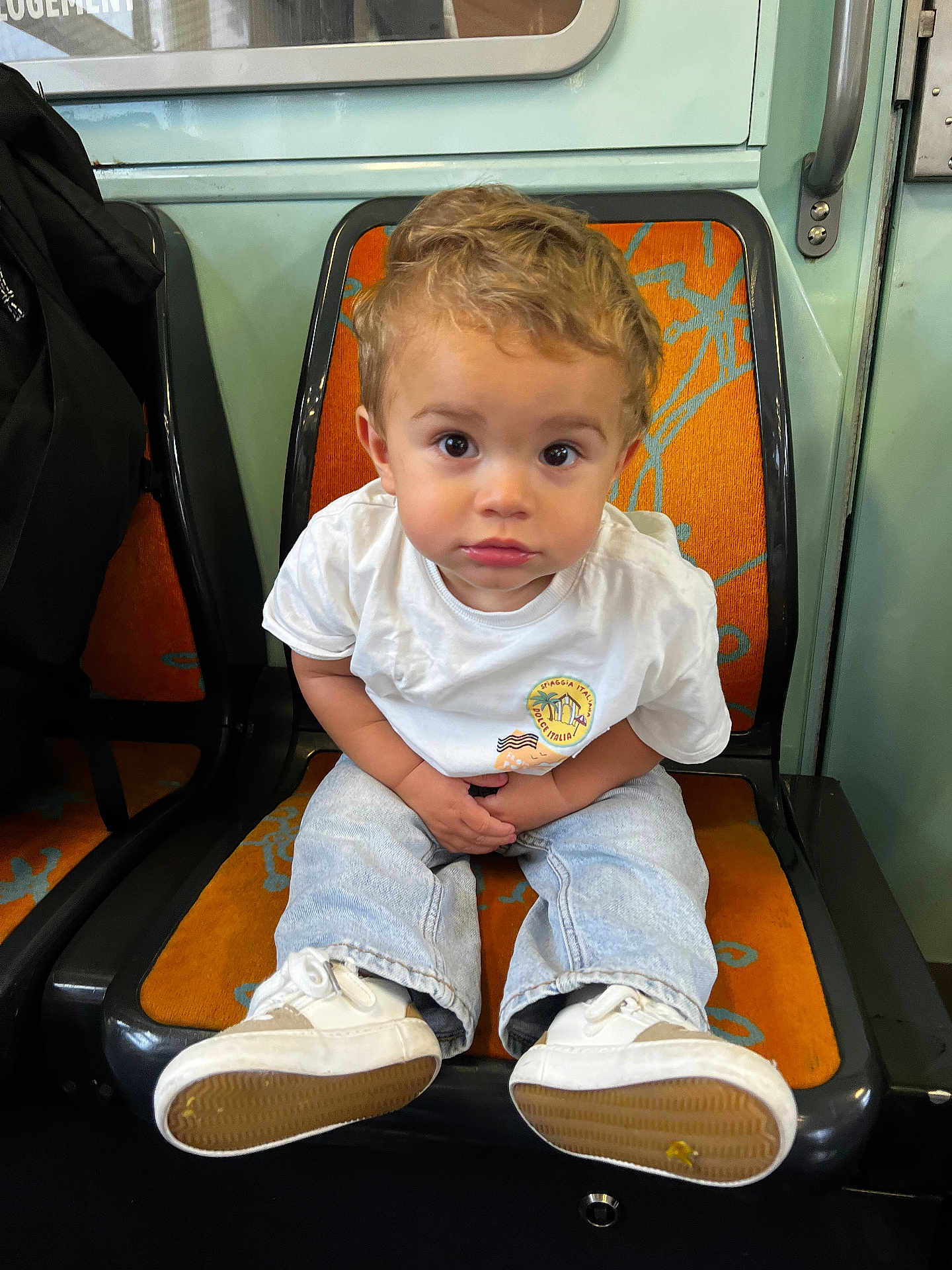 Louis participe au concours pour gagner de l'argent avec cette photo : toddler, child, sitting, white_tshirt, jeans, shoes, orange_seat, public_transport, curly_hair, indoor, looking_at_camera, young_child, casual_clothing, seat, floor, hands, expression, face, person, transport