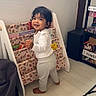 toddler, child, smiling, standing, barefoot, indoor, books, bookshelf, toy, stuffed_animal, basket, floor, window, natural_light, clothing, white_shirt, pants, furniture, room, person