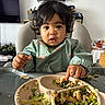 Meije a rejoint le concours — aidez-le/la à gagner de superbes lots ! toddler, child, high_chair, messy, food, peas, chicken, plate, dining, indoor, curly_hair, green_bib, eating, table, meal, portrait, baby_food, hands, face, cute