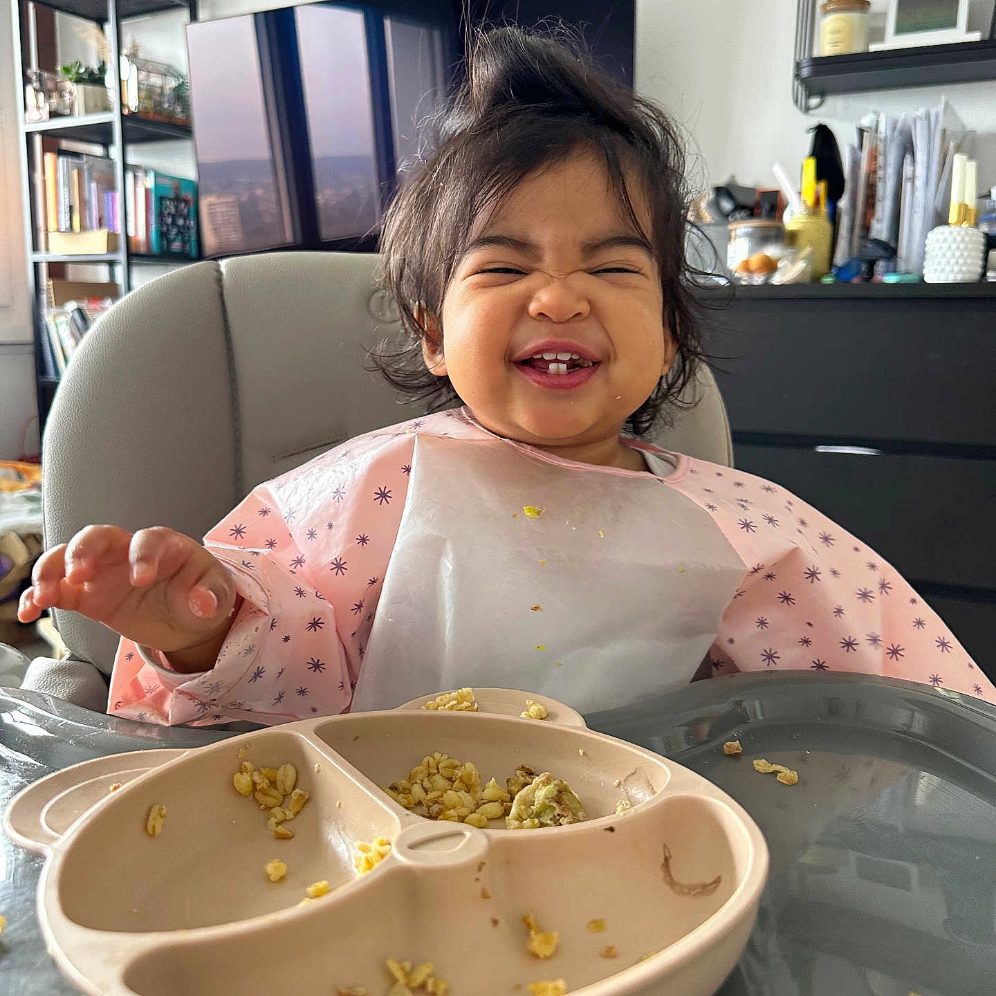 Meije participe au concours pour gagner de l'argent avec cette photo : toddler, child, high_chair, bib, food, plate, smile, happy, messy_hair, indoor, living_room, furniture, plant, shelf, book, table, person, face, clothing, home