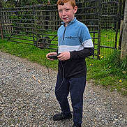 Jake is registered to the contest to win money with this photo: boy, casual_clothing, child, daylight, device, fence, grass, gravel, holding, nature, outdoor, pants, red_hair, shoes, sky, smiling, standing, sweatshirt, trees, wire