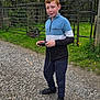 boy, casual_clothing, child, daylight, device, fence, grass, gravel, holding, nature, outdoor, pants, red_hair, shoes, sky, smiling, standing, sweatshirt, trees, wire