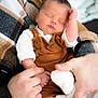 baby, newborn, sleeping, infant, hands, blanket, plaid_blanket, brown_overalls, white_shirt, cozy, portrait, closeup, soft_light, adult_hands, knitted_sock, skin, peaceful, indoor, parent, cuddling