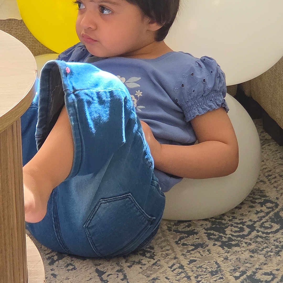 Izna joined the competition — help win amazing prizes! child, toddler, portrait, sitting, indoors, balloons, yellow_balloon, white_balloon, blue_clothing, jeans, chair, furniture, carpet, rug, table, sunlight, natural_light, brown_hair, looking_away, casual_photo