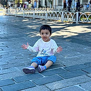 Izna joined the competition — help win amazing prizes! child, toddler, outdoor, street, pavement, sitting, portrait, smiling, expressive, hands_raised, short_hair, white_shirt, blue_pants, sneakers, city, sidewalk, railing, pedestrians, daylight, urban_scene