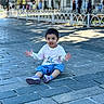 child, toddler, outdoor, street, pavement, sitting, portrait, smiling, expressive, hands_raised, short_hair, white_shirt, blue_pants, sneakers, city, sidewalk, railing, pedestrians, daylight, urban_scene