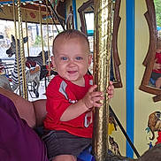 Cayden joined the competition — help win amazing prizes! child, toddler, red_shirt, carousel, golden_pole, amusement_ride, mirror, animal_figure, zebra, smiling, indoor, fun, playful, holding, shorts, blue_seat, reflection, person, family, entertainment