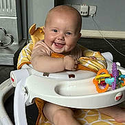 Lula is registered to the contest to win money with this photo: arm, baby, baby_walker, child, clothing, face, floor, furniture, happy, head, home, indoor, leg, metal_decoration, padding, power_outlet, smiling, toy, wall, yellow