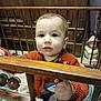 toddler, child, crib, wooden_bars, orange_sweater, penguin_design, indoor, furniture, baby, hand, face, person, curious, standing, home, coat, young_child, portrait, expression, room