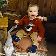 Charli participe au concours pour gagner de l'argent avec cette photo : baby, blue_socks, child, christmas_tree, cushions, decor, festive, floor_tiles, gnome, hand, holiday, indoor, pants, person, red_clothing, smiling, sweater, toddler, toy, wooden_door