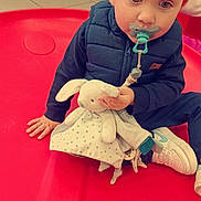 Tiago participe au concours pour gagner de l'argent avec cette photo : toddler, child, pacifier, toy, stuffed_animal, bunny, blue_vest, sneakers, red_play_surface, indoor, floor_tiles, curious, sitting, hand, face, blond_hair, clothing, young_child, play_area, comfort