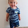 toddler, child, boy, smiling, standing, indoor, casual_clothing, blue_shirt, shorts, puma, footwear, crocs, colorful, floor, wooden_floor, wall, cute, happy, portrait, playful