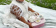 Autumn is registered to the contest to win money with this photo: baby, child, white_dress, lace, feathers, outdoor, grass, purse, pearls, flowers, head_accessory, cute, sitting, portrait, nature, greenery, yard, house, pretty, expression