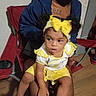 toddler, child, adult, yellow_clothing, headband, bow, sitting, lap, chair, indoor, wooden_floor, blue_hoodie, red_chair, person, curly_hair, casual, portrait, cozy, family, candid