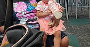 Jream is registered to the contest to win money with this photo: child, toddler, woman, indoor, bottle, pink_clothing, headband, curly_hair, sitting, chairs, play_area, carpet, sneakers, bag, holding, person, expression, casual_clothing, floor, stroller