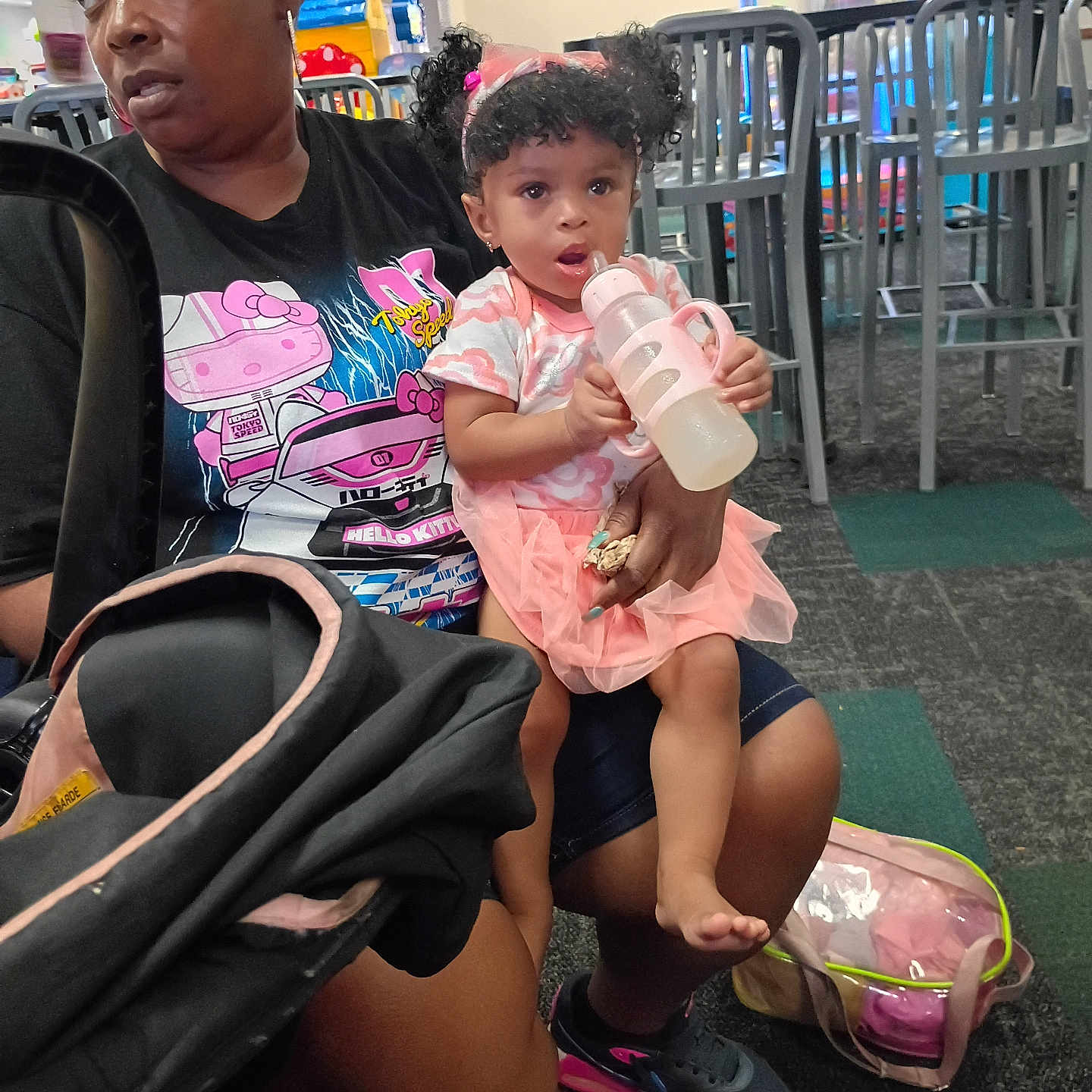 Jream is registered to the contest to win money with this photo: child, toddler, woman, indoor, bottle, pink_clothing, headband, curly_hair, sitting, chairs, play_area, carpet, sneakers, bag, holding, person, expression, casual_clothing, floor, stroller