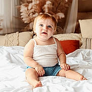 Orianne a rejoint le concours — aidez-le/la à gagner de superbes lots ! toddler, child, baby, sitting, bed, white_bedspread, blue_eyes, tank_top, denim_shorts, indoor, pillows, cozy, decor, home, portrait, cute, smiling, blond_hair, soft_light, relaxed