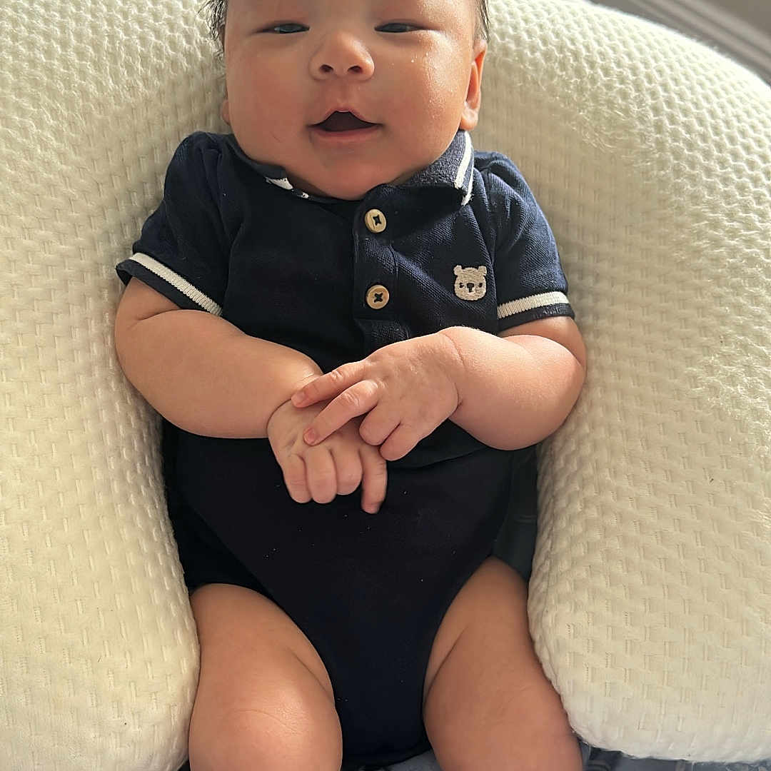 Zyron is registered to the contest to win money with this photo: baby, bear_emblem, bed, blanket, child, comfort, cozy, cushion, face, hands, indoors, infant, navy_blue, onesie, portrait, resting, skin, smile, soft_light, young_child