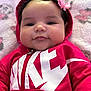 Nova is registered to the contest to win money with this photo: baby, infant, hoodie, red_clothing, pink_bow, curly_hair, face, cheeks, blanket, patterned_blanket, soft_texture, child, portrait, cute, smiling, indoor, clothing, person, relaxing, young_child