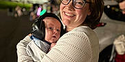 Jaxon is registered to the contest to win money with this photo: woman, baby, glasses, earmuffs, night, outdoor, smile, person, holding, happy, protective_gear, jacket, streetlights, people, blurred_background, parent, infant, embrace, warmth, candid